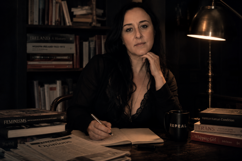 Cinematic portrait of Sorcha Rosa seated at a laptop in a softly lit study surrounded by books, representing political writing, cultural commentary, feminism and modern Irish public life.
